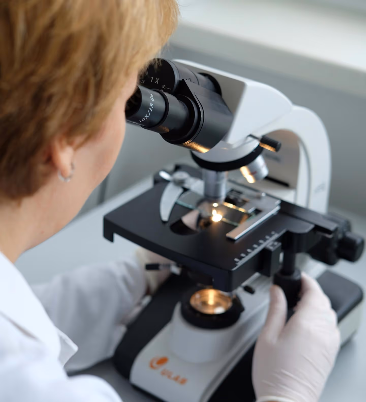 Scientist with blonde hair wearing white gloves observing a sample through a microscope.