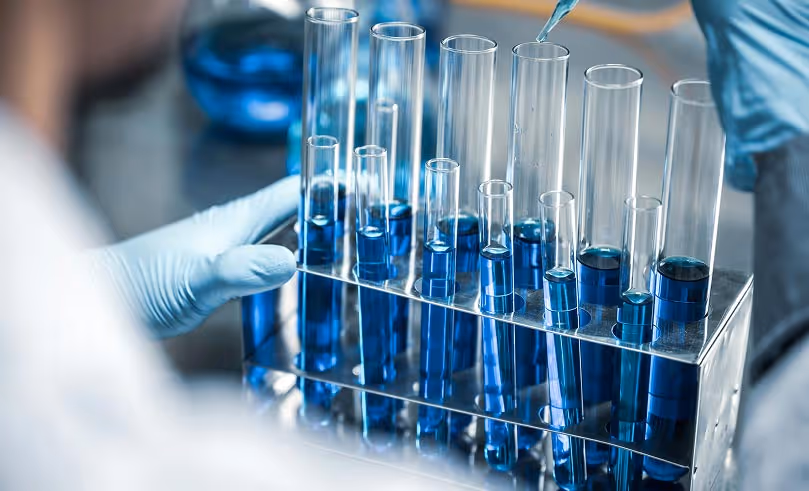 Scientist wearing gloves holding a rack of test tubes filled with varying levels of blue liquid while adding liquid with a dropper.