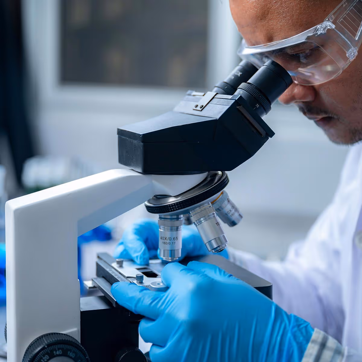 Scientist wearing safety goggles and blue gloves adjusting a microscope slide in a laboratory.